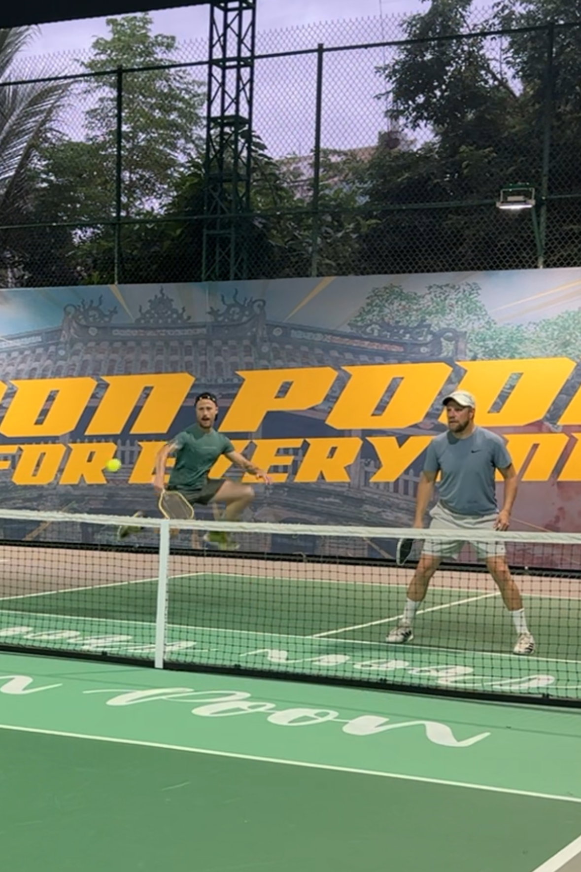 Two people playing on a covered pickleball court with a large banner in the background.