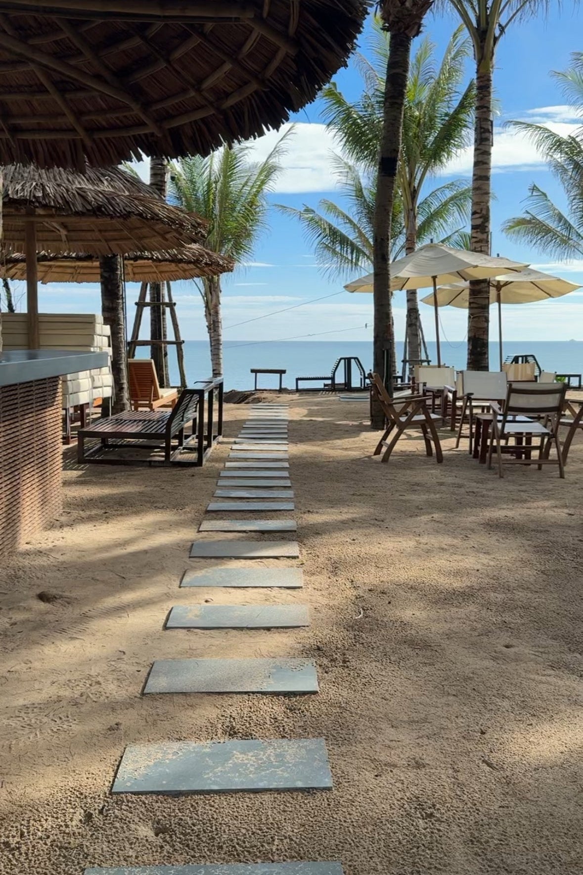 Beachside dining area with tables, chairs, and umbrellas under palm trees.