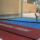 Child playing pickleball on a court with a red and blue surface and palm trees in the background.