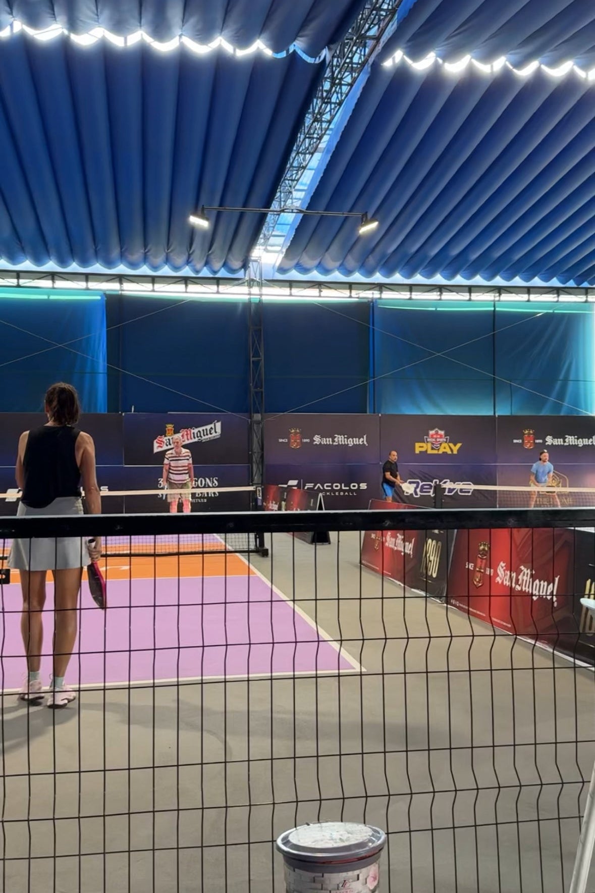 Indoor sports facility with people playing pickleball, blue roof, and promotional banners.