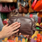Person holding a brown leather dopp kit in front of a colorful display of bags and shoes.