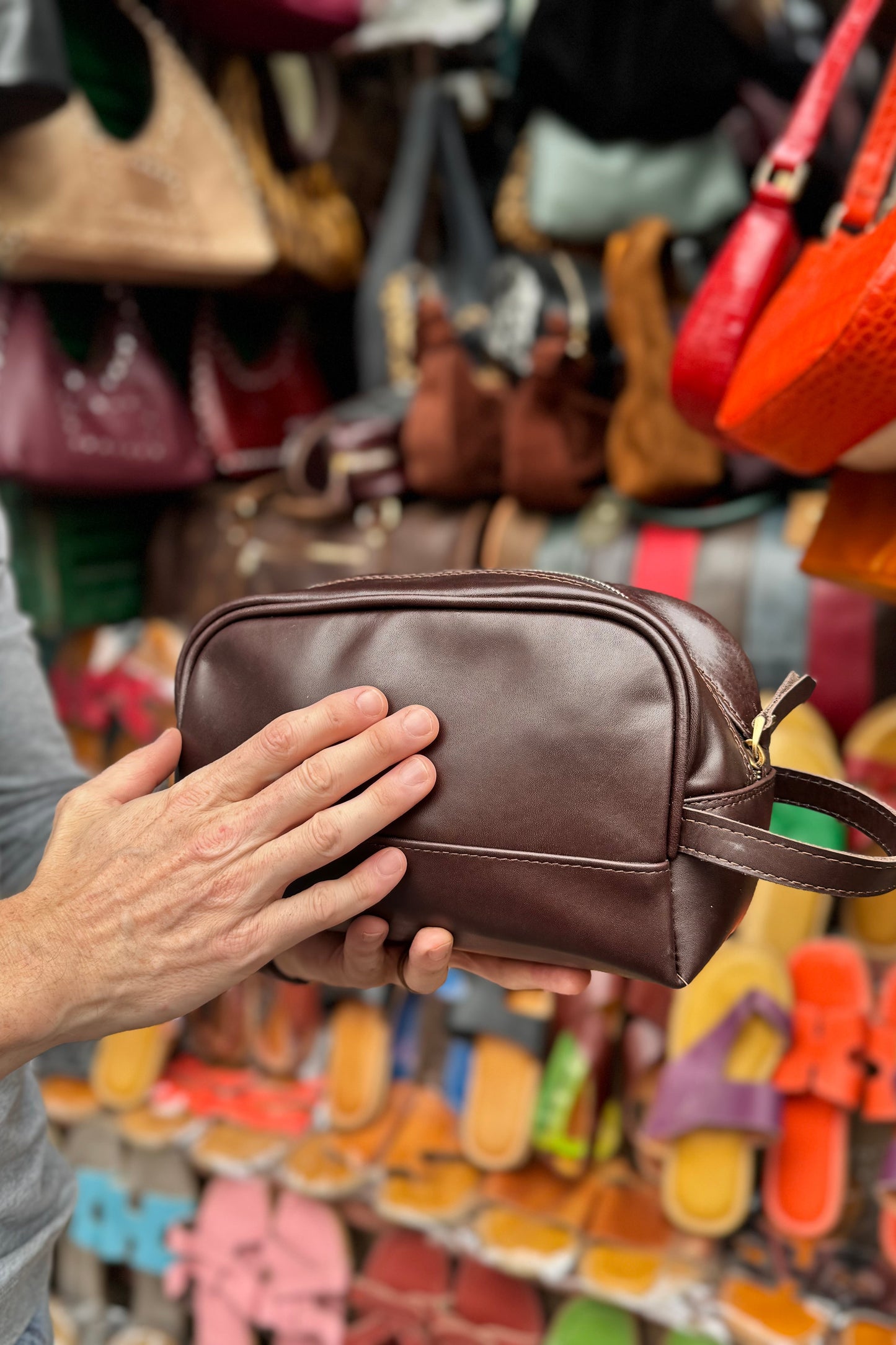 Person holding a brown leather dopp kit in front of a colorful display of bags and shoes.