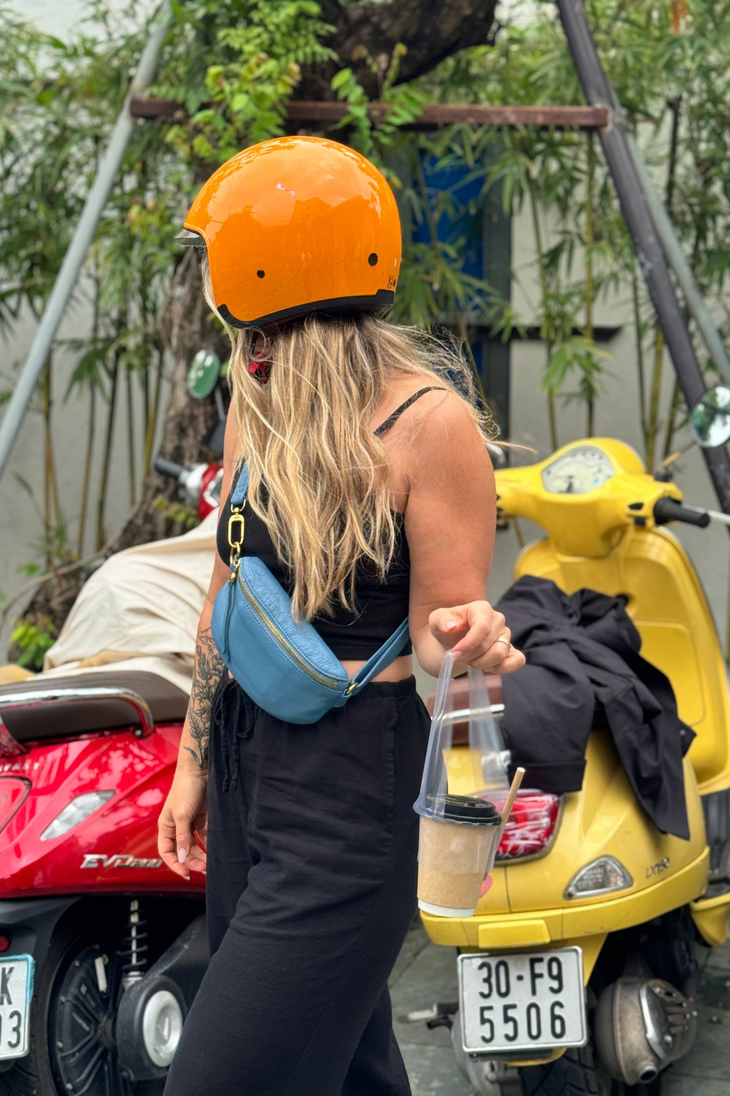 Person wearing an orange helmet and black outfit with a blue bag, standing next to a yellow scooter.