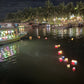 Decorative lights floating on water with boats and palm trees in the background at night.