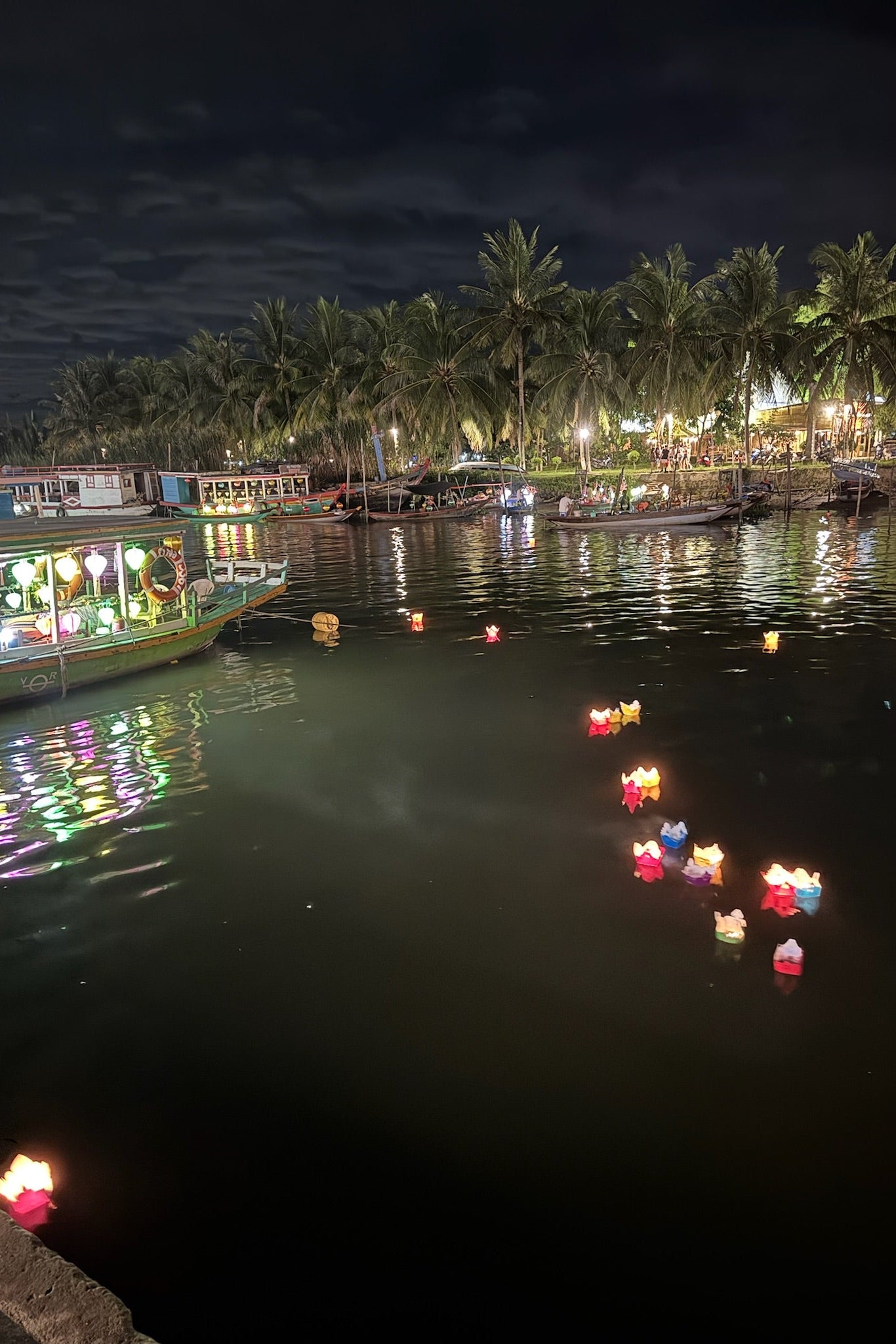 Decorative lights floating on water with boats and palm trees in the background at night.