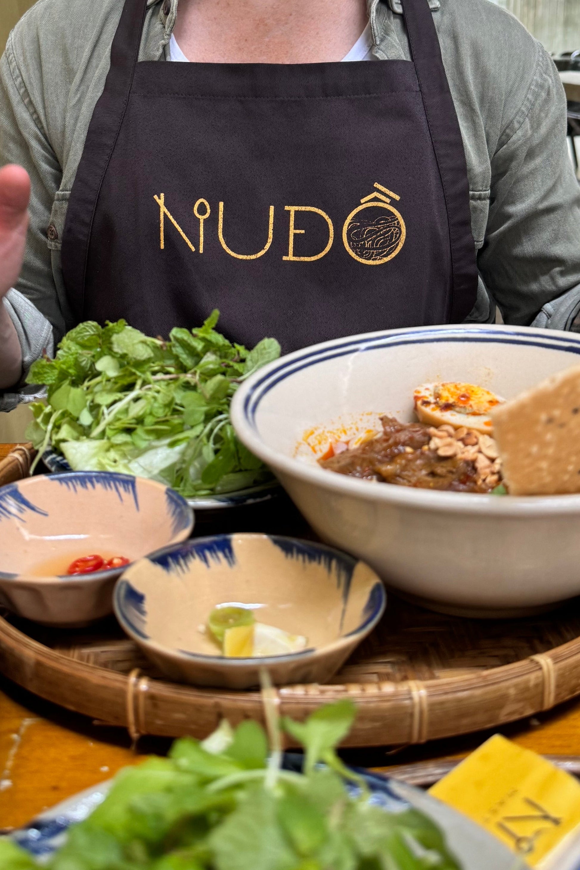 Man in a kitchen wearing a 'Nudo' apron, surrounded by food items.