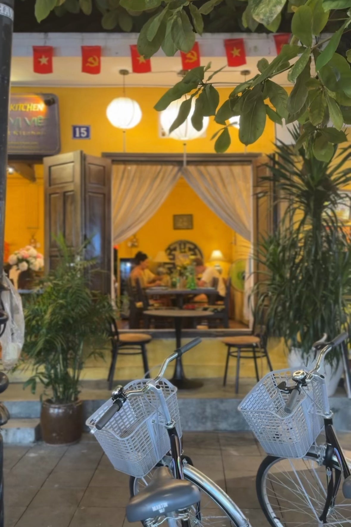 Outdoor seating area with bicycles, plants, and a yellow building interior.