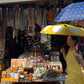 Woman holding a yellow umbrella over a market stall with various items on display.