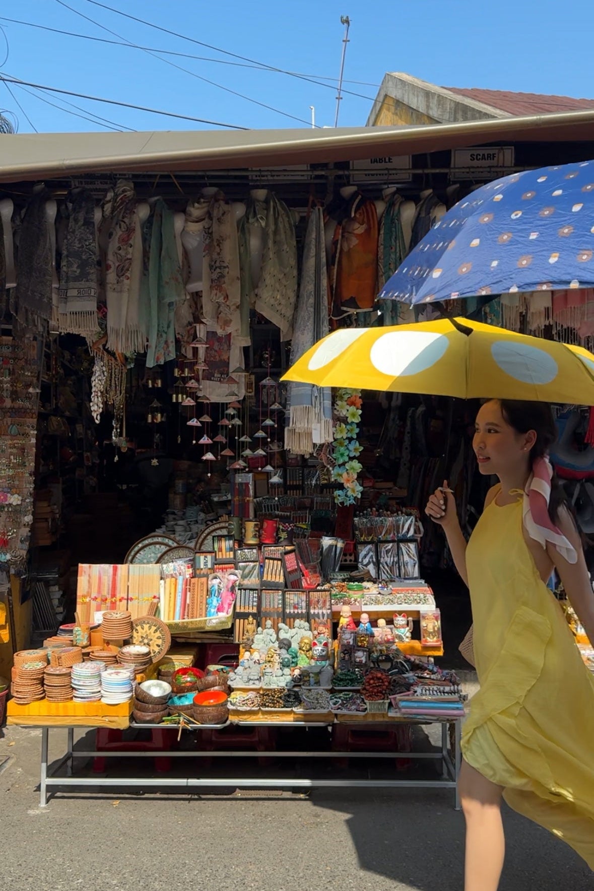 Woman holding a yellow umbrella over a market stall with various items on display.