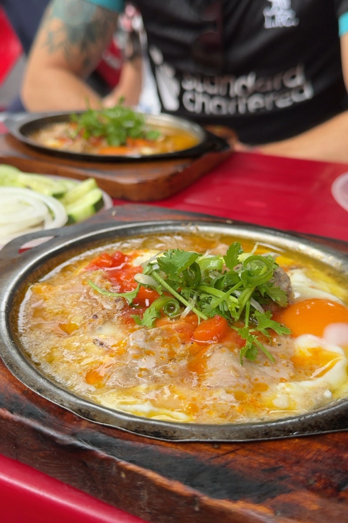 Hot dish with vegetables and eggs on a wooden table, person in black shirt in the background