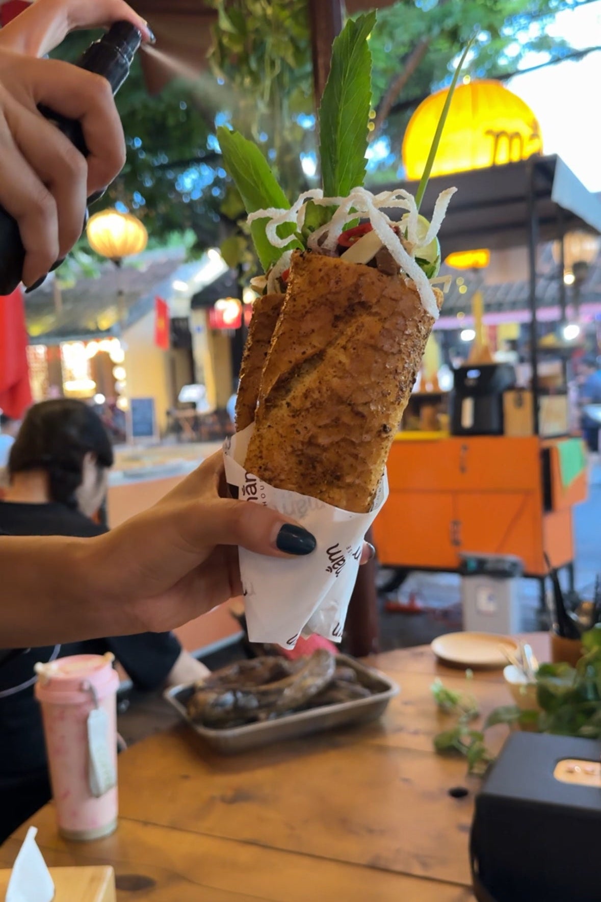 Person holding a crispy banh mi with green leaves in an outdoor setting.